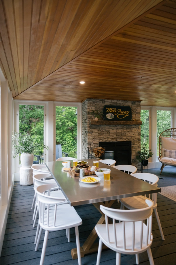 Indoor patio dining space with long table, white chairs, stone fireplace, and glass walls opening to surrounding greenery
